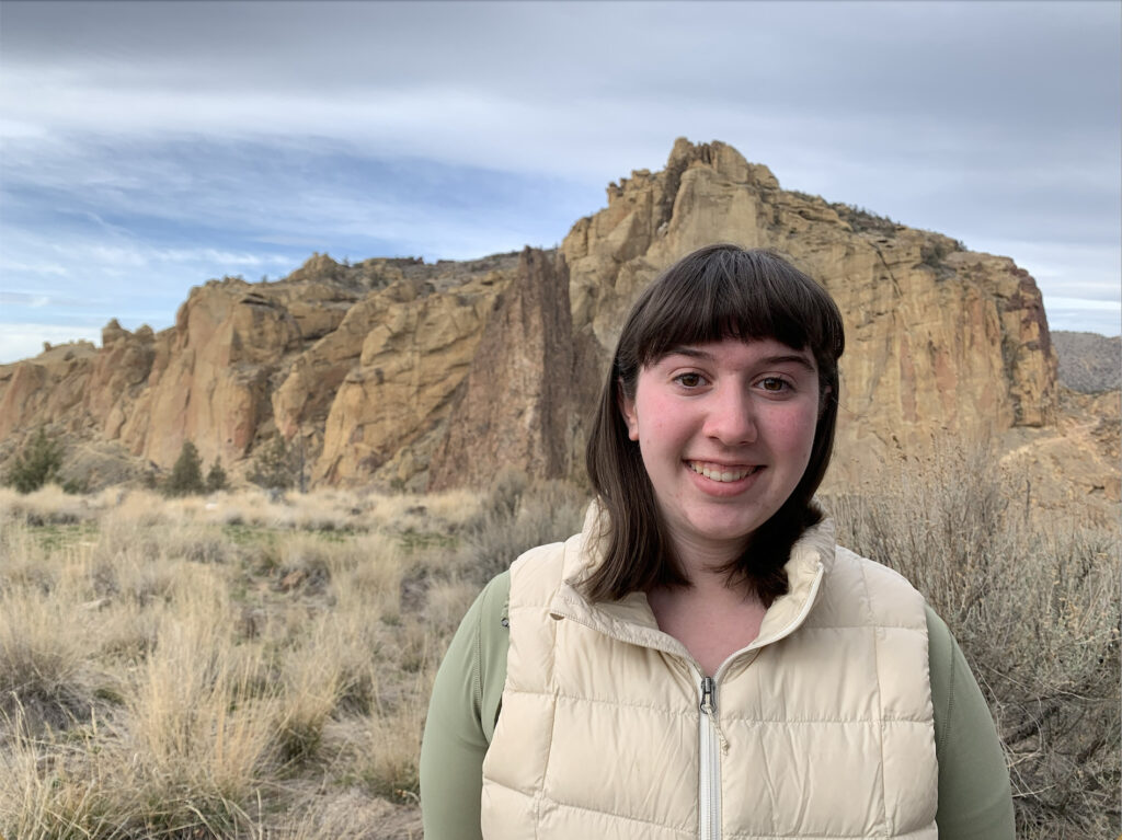 a headshot of a person with dark shoulder length hair and bangs standing in front of a beautiful background of Smith Rocks