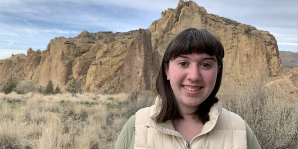 a headshot of a person with dark shoulder length hair and bangs standing in front of a beautiful background of Smith Rocks
