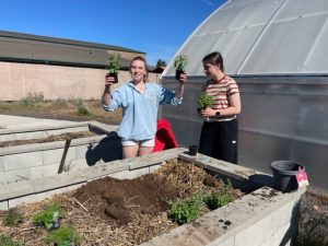 two high school girls hold plants next to a garden bed