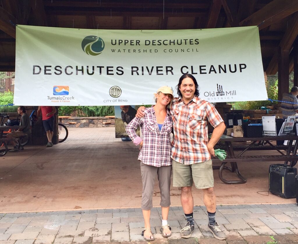two people stand together smiling in front of a sign that reads Deschutes River Cleanup