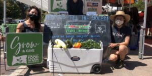 A portable cooler full of fresh produce at an outdoor booth with three people around it