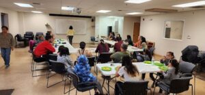 Youth and adults sitting at tables with green reusable plates in a community room