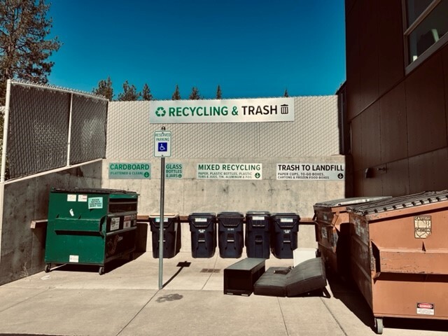 Trash and recycling dumpsters and carts with large metal signage outside COCC's Wickiup residence hall