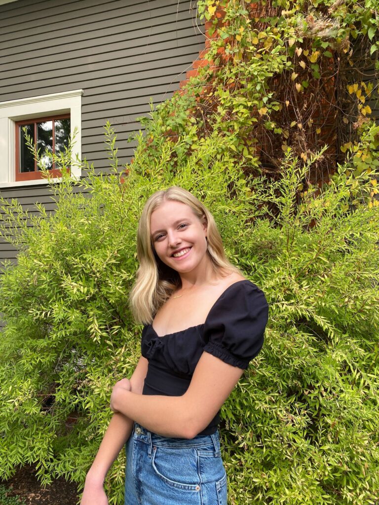 a young woman with blonde hair and light skin poses smiling in front of a background of green foliage. she is wearing a black short sleeve top.