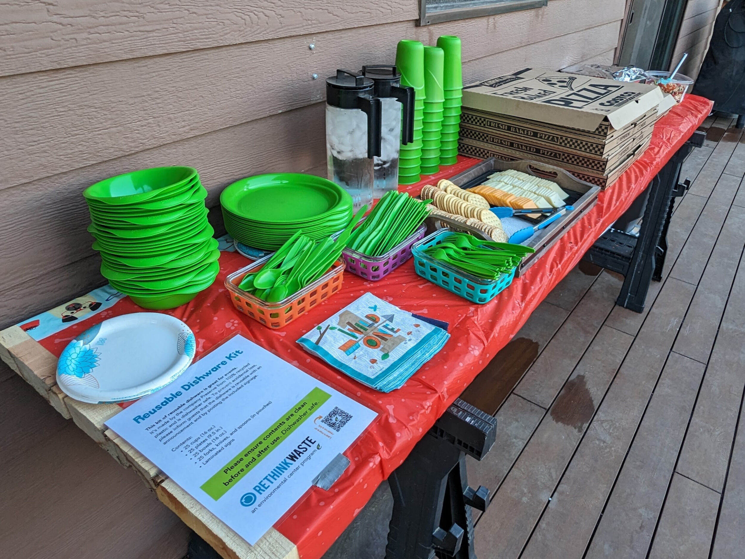 a long table set up for a birthday party with green reusable dishware. there are pizza boxes and cheese and crackers on the table.
