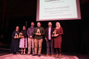Six people stand on a stage smiling. Four are holding wooden awards. A screen behind them references the Sustainability Awards.