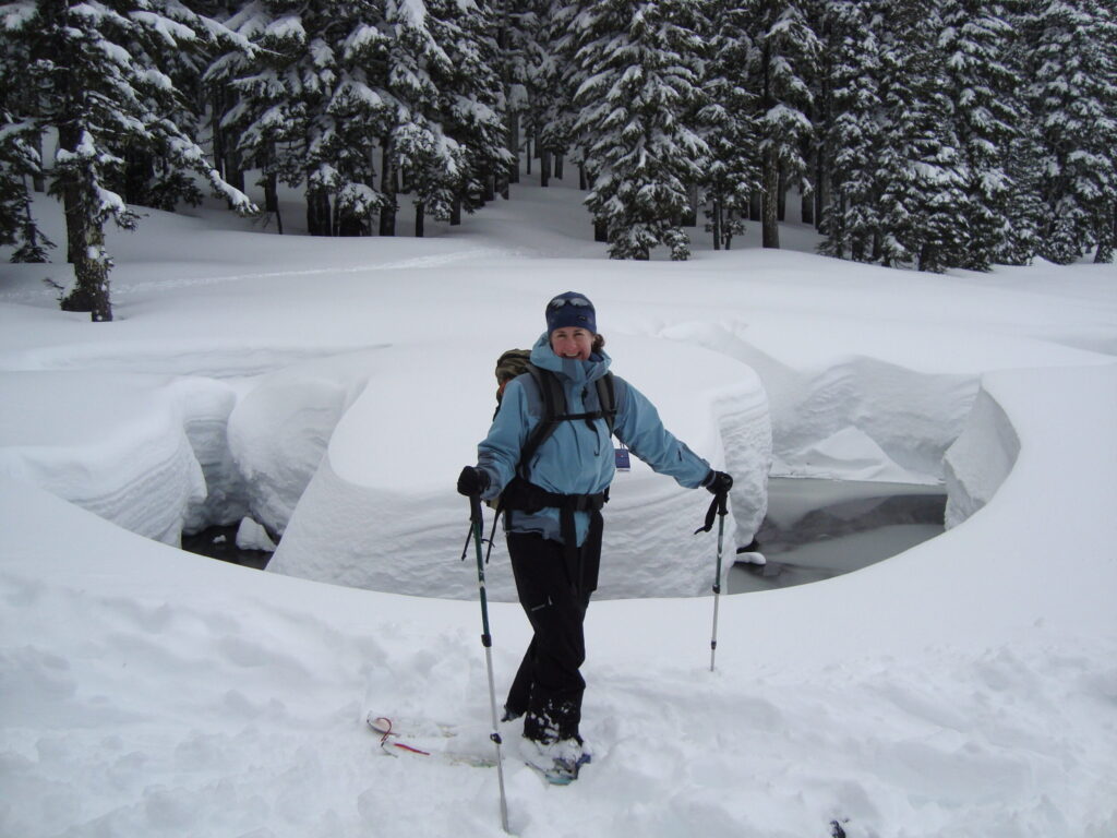 a woman in a ski hat, light blue ski jacket, black ski pants and a backpack smiles in the snow. she is wearing snowshoes and holding ski poles.