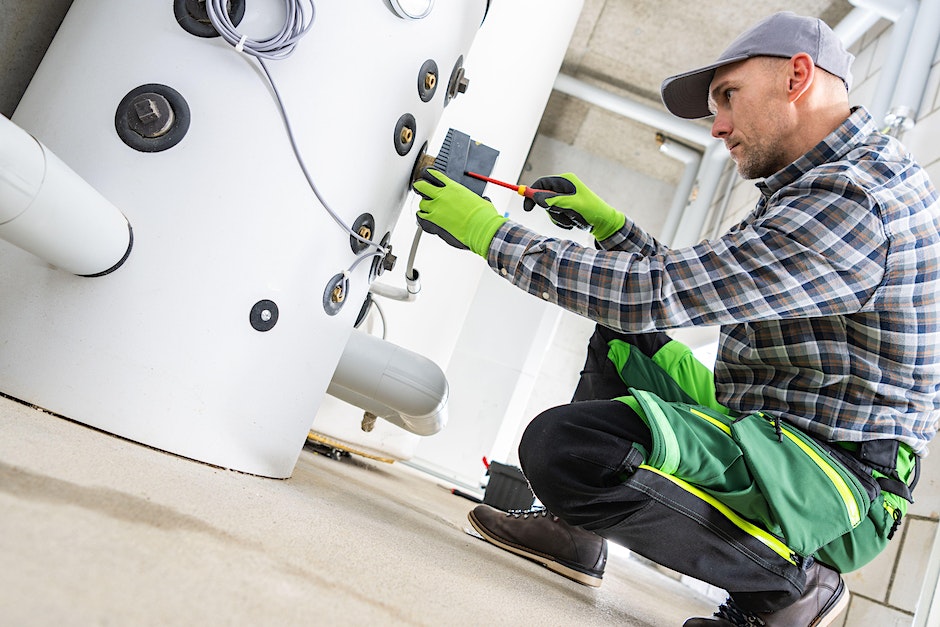 a man wearing a plaid shirt, green work gloves, and a baseball cap kneels to fix a water heater with tools