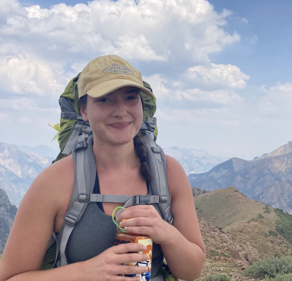 a young woman wearing a tan baseball cap and a green backpacking pack stands in front of a mountain view, smiling