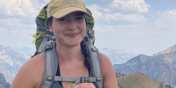 a young woman wearing a tan baseball cap and a green backpacking pack stands in front of a mountain view, smiling