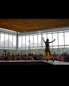 A model on stage in the foreground with a crowd of people in the background of a window-paneled arena