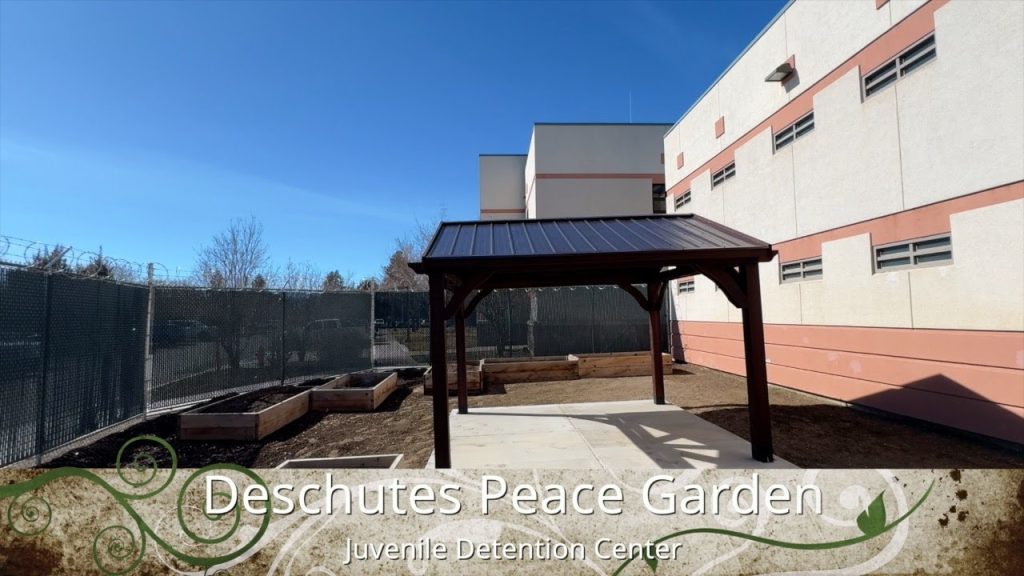Fenced garden with plant beds and a shade structure with solar panel with this text overlaid at the bottom: "Deschutes Peace Garden - Juvenille Detention Center"