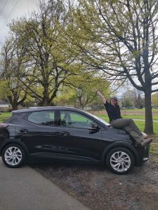 a young woman wearing a black tshirt and green pants sits on the hood of a black electric vehile. she is posing with a smile and her hand in the air