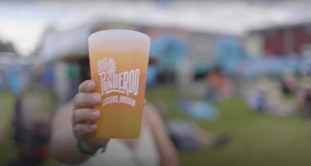 A person holding a Big Ponderoo silicone pint glass with the festival out of focus in the background