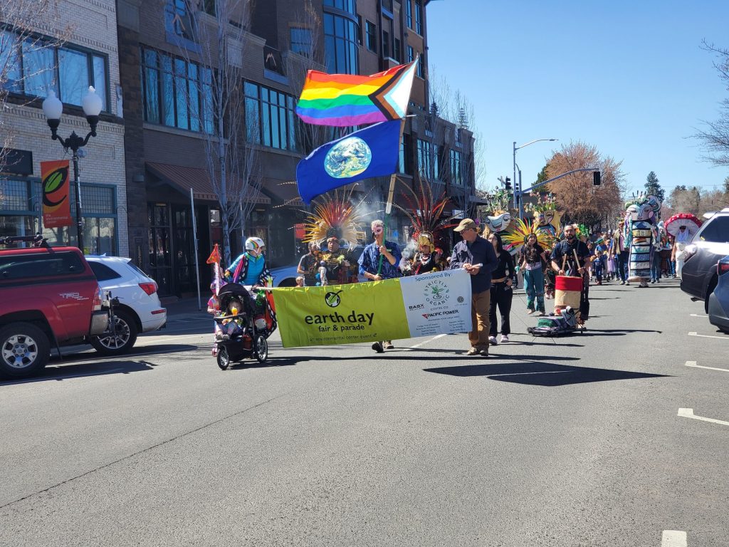 a parade travels down a street in Bend Oregon. the leader of the parade holds a flagpole with an earth flag and a LGBTQ+ pride flag, as well as a banner reading "earth day fair and parade"