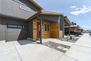 new housing development with multiplex-style homes. homes are grey with wood accents and red doors, showing clean driveways and a few cars in the distance parked at home entrances.