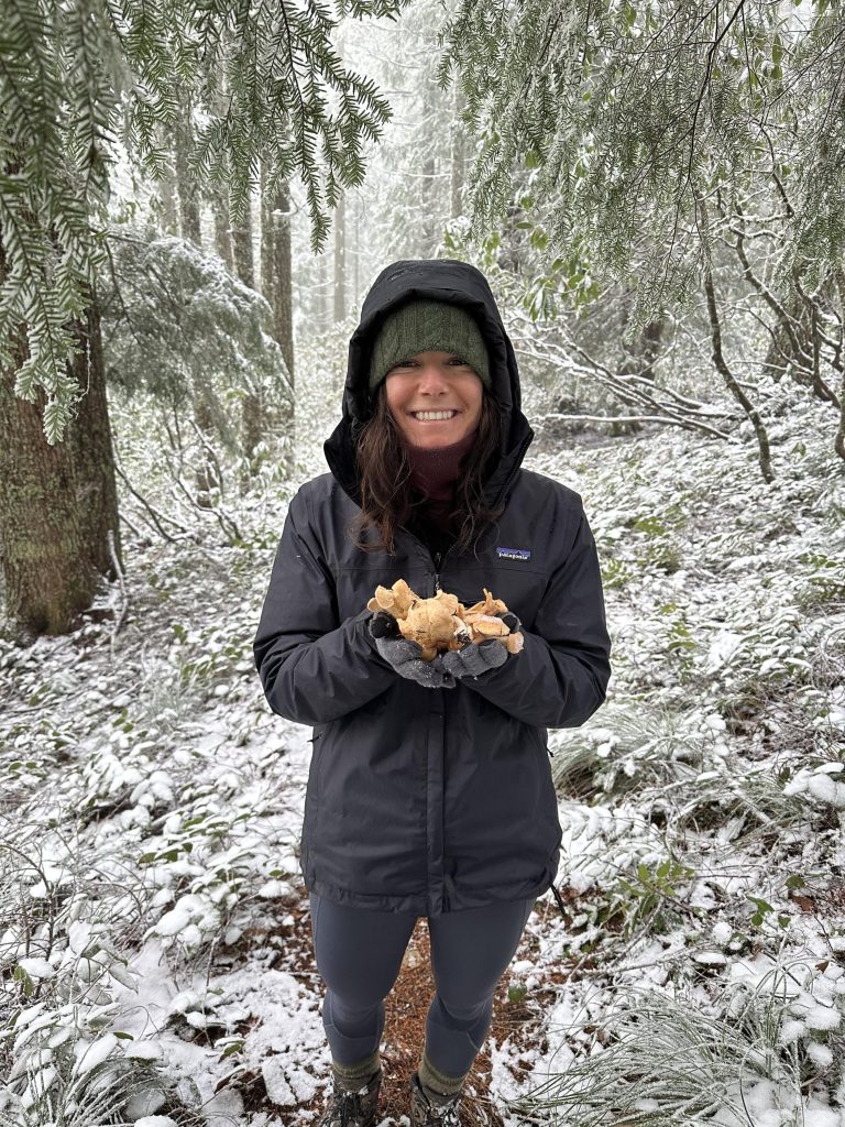 a woman wearing a black hooded rain jacket and black pants smiles and poses. she is holding wild mushrooms that she foraged. she is in a snowy forest.