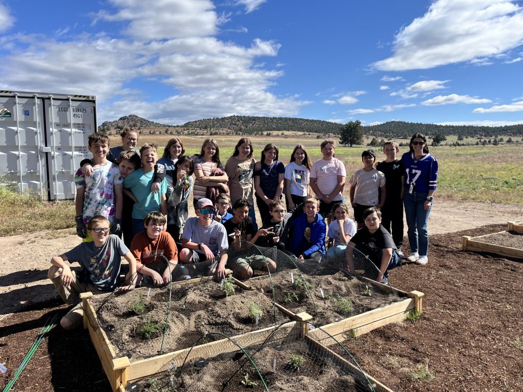 a group of 5th graders and their teachers pose in front of new garden beds in their school yard. behind them the corner of the school is visible and a blue sky with white clouds.