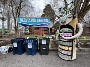 recycling bins under a banner, next to a tall puppet creature