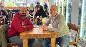 Two men sitting across from each other at a table in a restaurant looking at the camera smiling