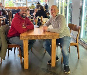 Two men sitting across from each other at a table in a restaurant looking at the camera smiling