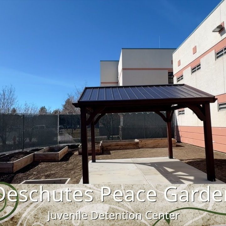 Fenced garden with plant beds and a shade structure with solar panel with this text overlaid at the bottom: "Deschutes Peace Garden - Juvenille Detention Center"