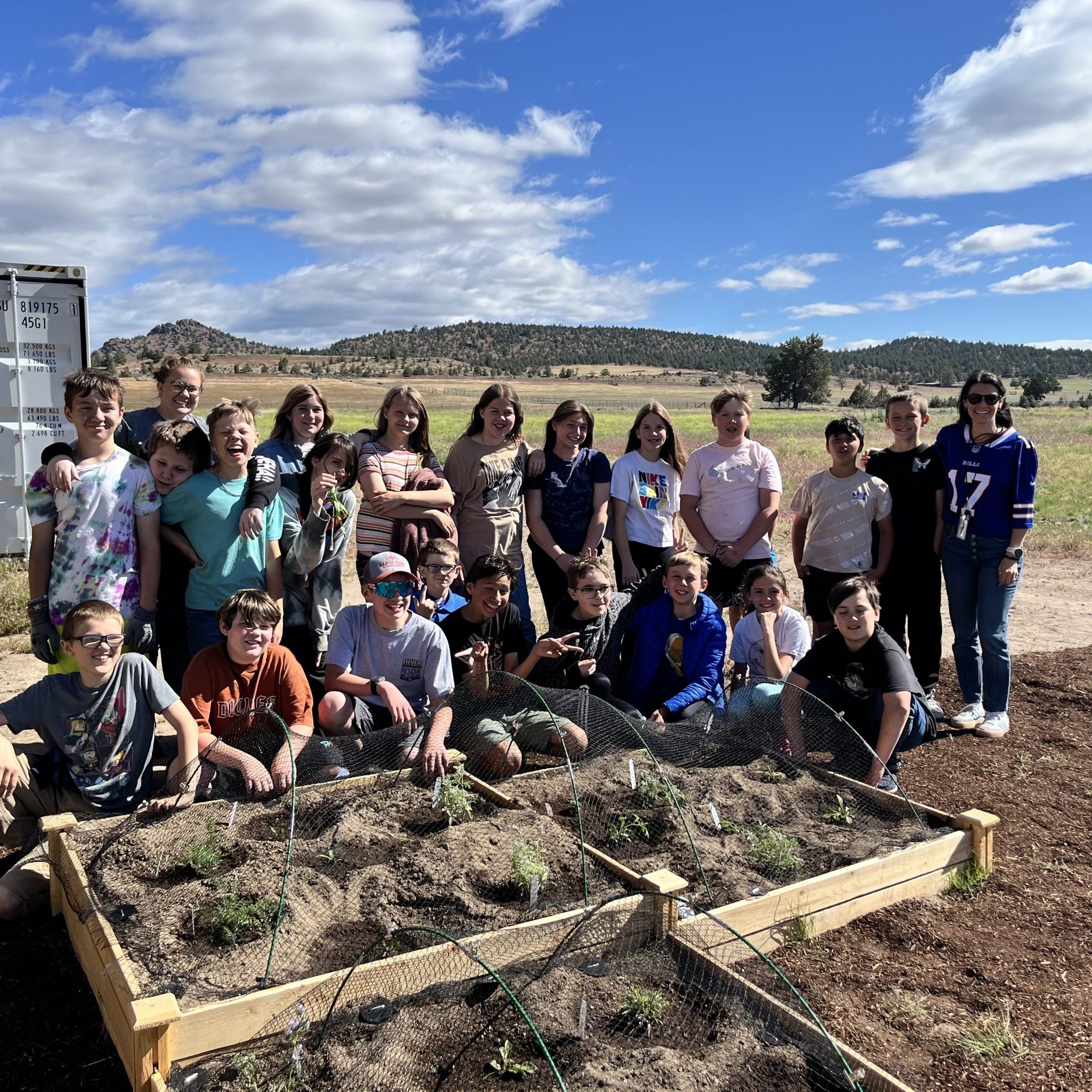 a group of 5th graders and their teachers pose in front of new garden beds in their school yard. behind them the corner of the school is visible and a blue sky with white clouds.