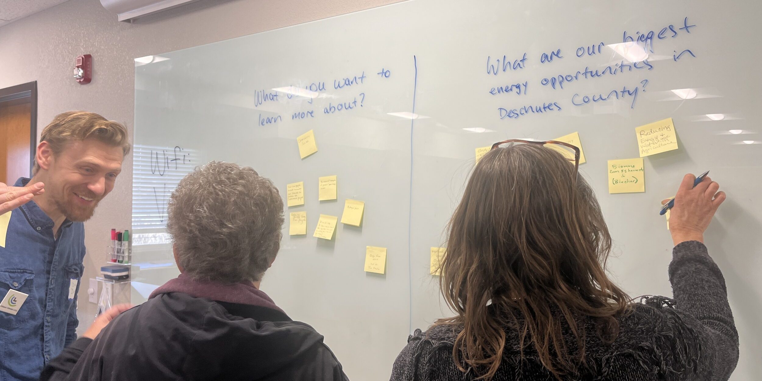 Three people gathered around a whiteboard, talking and putting yellow sticky notes in categories