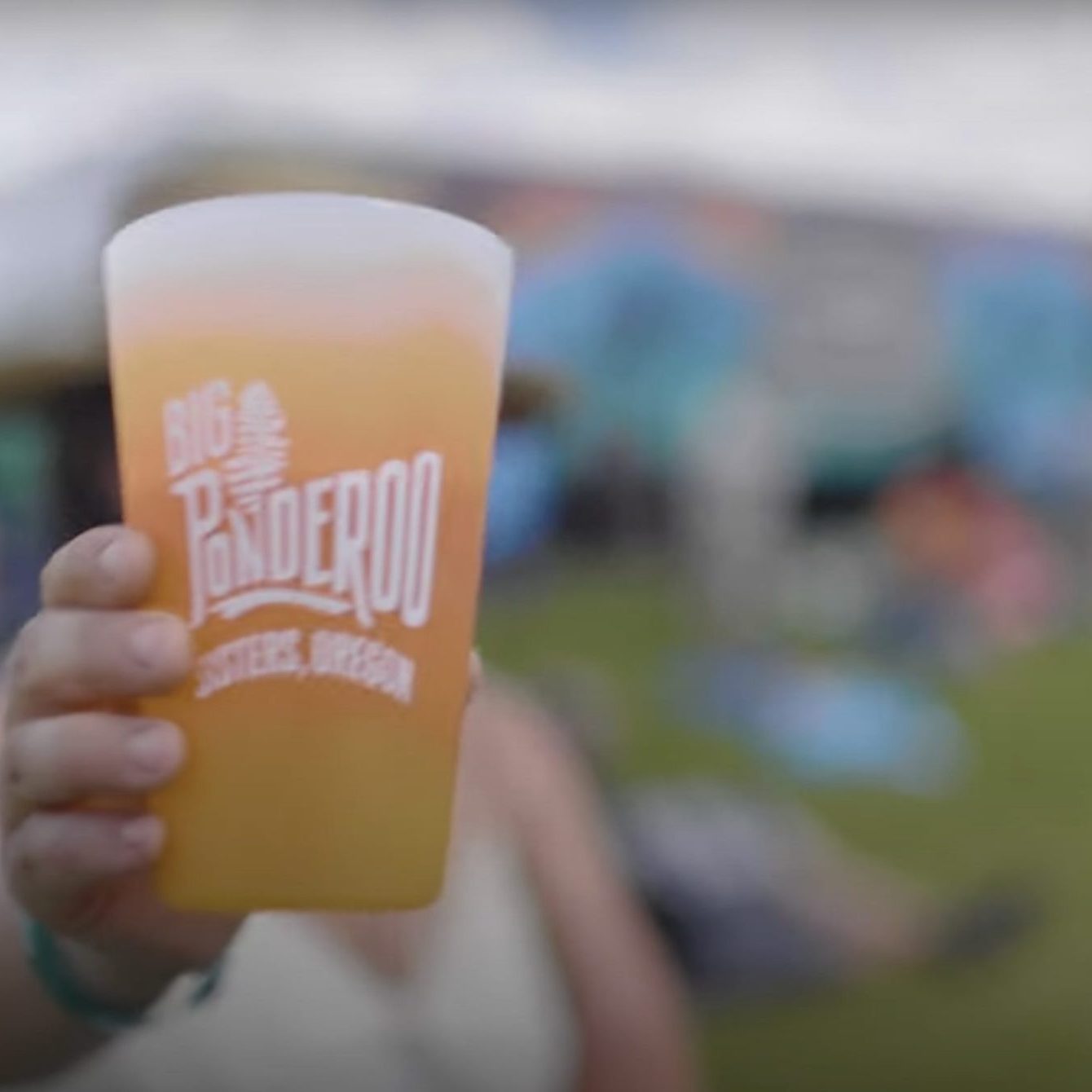 A person holding a Big Ponderoo silicone pint glass with the festival out of focus in the background