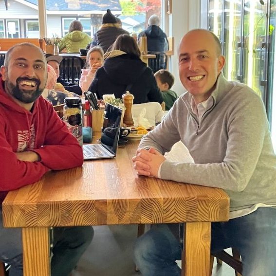 Two men sitting across from each other at a table in a restaurant looking at the camera smiling